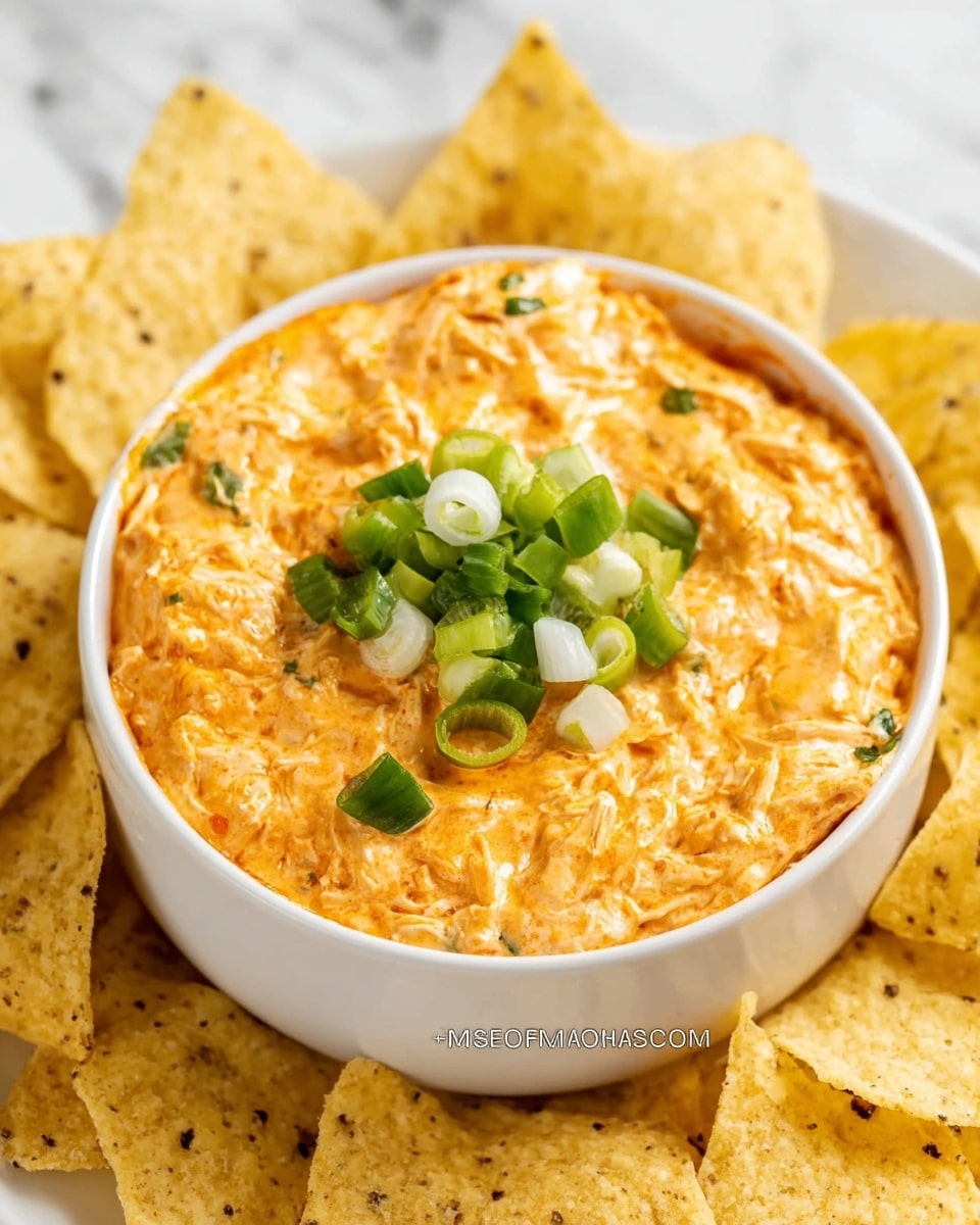 A white bowl filled with a creamy, orange buffalo chicken dip that has a slightly rough texture with visible shredded chicken pieces mixed throughout; on top of the dip is a small heap of chopped green onions in different shades of green and white, adding a fresh contrast; around the bowl, there are light yellow corn tortilla chips with a grainy surface and some specks, arranged to frame the bowl on a white marbled texture background. photo taken with an iphone --ar 4:5 --v 7