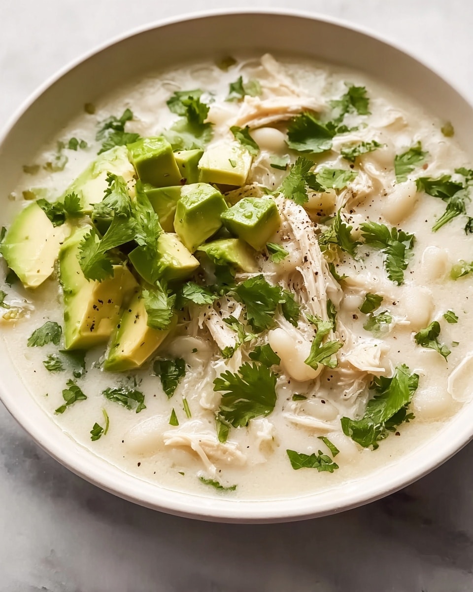 A white bowl filled with creamy white soup that has shredded chicken and soft white hominy as the base layer, garnished with bright green chopped cilantro scattered throughout. On top, there are chunks of pale green avocado pieces adding a fresh color contrast, with extra cilantro leaves placed beside them. The soup looks thick and textured with a bit of black pepper sprinkled lightly, all placed on a white marbled surface. photo taken with an iphone --ar 4:5 --v 7
