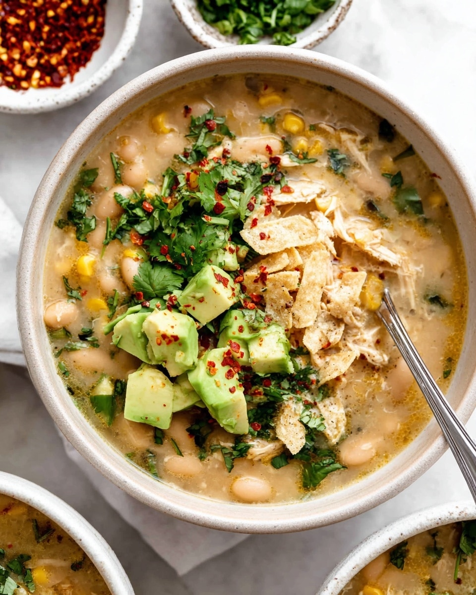 A bowl of creamy white bean soup with shredded chicken and pieces of corn fills most of the bowl, showing a light beige broth with a slightly thick texture. On top, there is a layer of diced bright green avocado chunks, sprinkled with red chili flakes and fresh chopped dark green cilantro leaves. Small broken pieces of light-colored tortilla chips are scattered over the top, adding texture. A silver spoon rests inside the bowl, placed near the avocado, all set on a white marbled surface. In the background, small white bowls hold red chili flakes and fresh parsley. Photo taken with an iphone --ar 4:5 --v 7