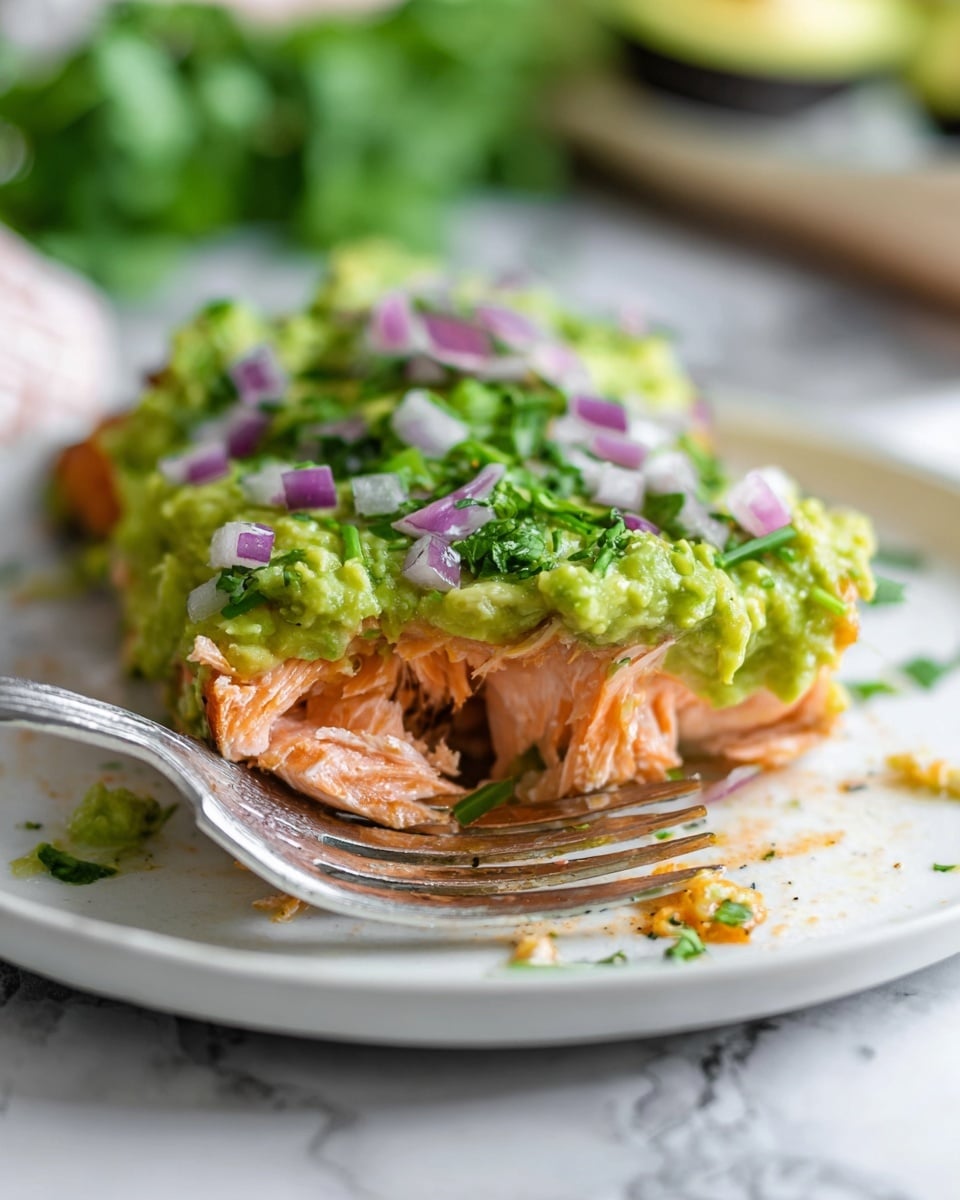 Two pieces of cooked salmon with a soft orange color and light grill marks lie side by side on a white plate. On top of each salmon piece is a thick layer of bright green mashed avocado, slightly chunky in texture. Scattered over the avocado are small cubes of red onion, purple and white in color, along with small sprigs of fresh green dill. The plate sits on a white marbled surface, with blurred background elements that include a bowl with more onions and lemons. Photo taken with an iphone --ar 4:5 --v 7