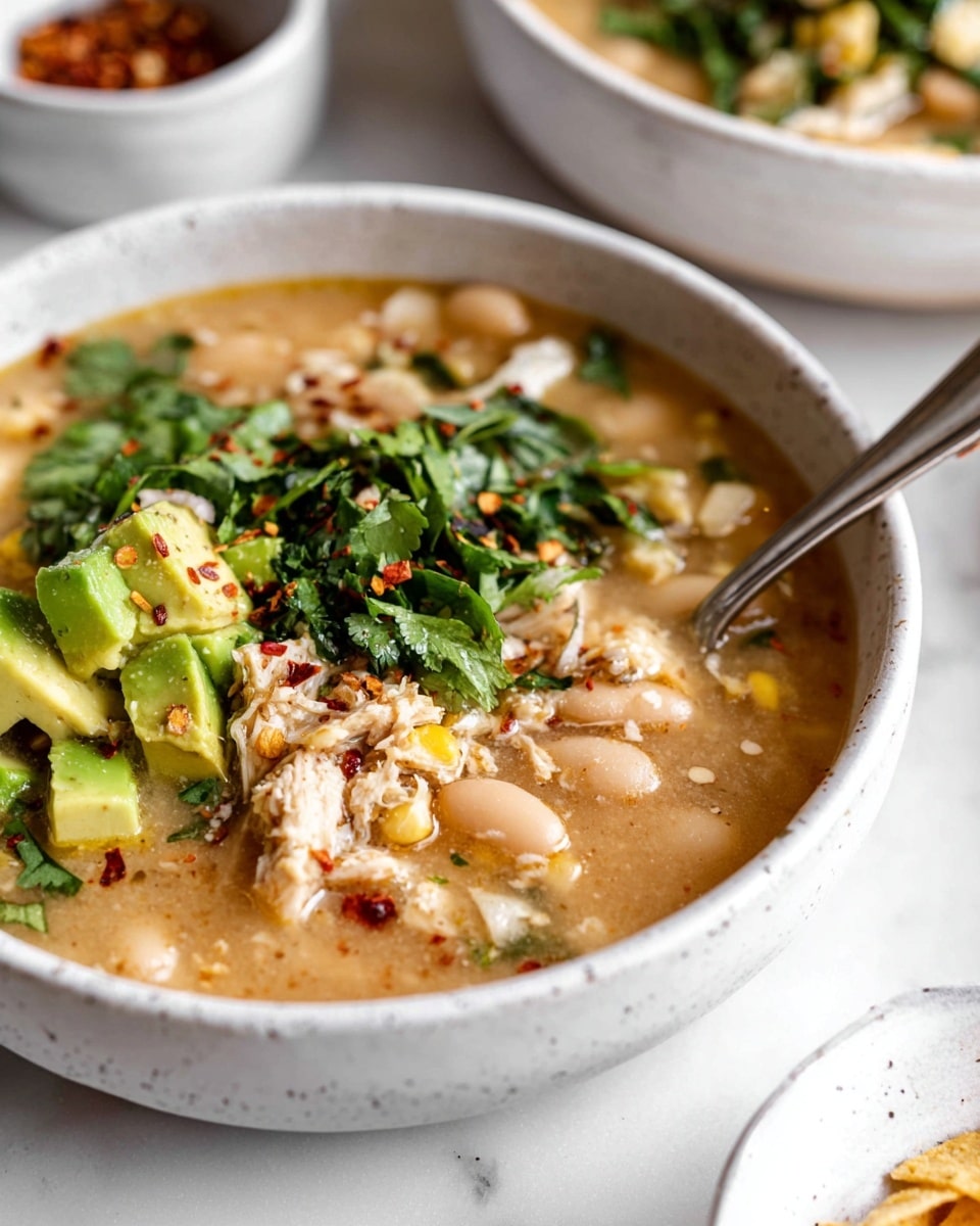A close-up view of a bowl filled with a creamy, light brown soup with white beans, soft corn pieces, and bits of chicken. On top, there are bright green chopped avocado chunks, fresh leafy green cilantro, and small pieces of crushed tortilla chips with red chili flakes sprinkled over everything. The bowl is white with a slight speckled texture and sits on a white marbled surface. In the background, a silver spoon rests inside the bowl, and there are blurred white bowls with toppings nearby. Photo taken with an iphone --ar 4:5 --v 7