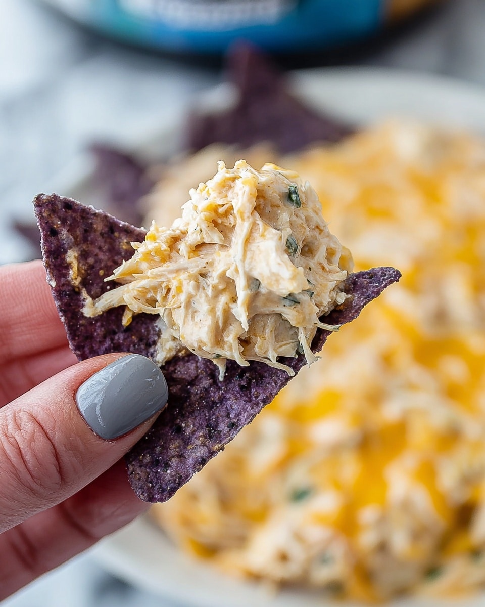 A close-up shows a purple tortilla chip held by a woman's hand with gray painted nails. The chip holds a scoop of creamy, light beige shredded chicken mixed with a smooth, cheesy white sauce, showing some small green herb flecks. In the blurred background, a white plate with a mix of melted white and yellow cheese is visible sitting on a white marbled surface. Photo taken with an iphone --ar 4:5 --v 7
