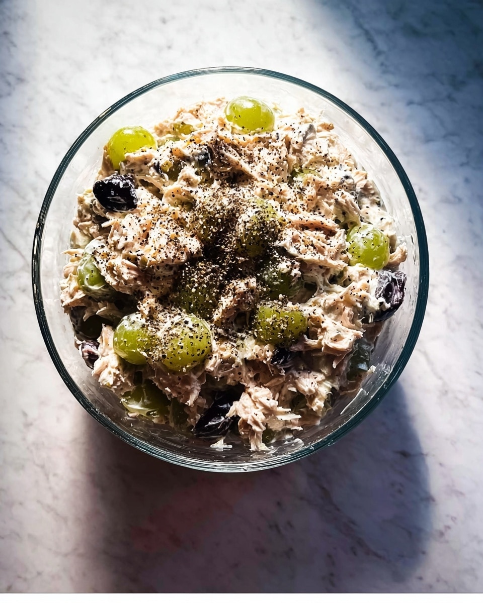 A clear glass bowl filled with a mixed tuna salad that shows about two main layers; the base layer is chunky tuna mixed with light creamy dressing, and scattered throughout are pieces of green grapes and dark bits like olives or black grapes. The top layer is sprinkled with coarse black pepper, adding texture and contrast. The bowl sits on a white marbled surface, and soft light casts gentle shadows around it. Photo taken with an iphone --ar 4:5 --v 7