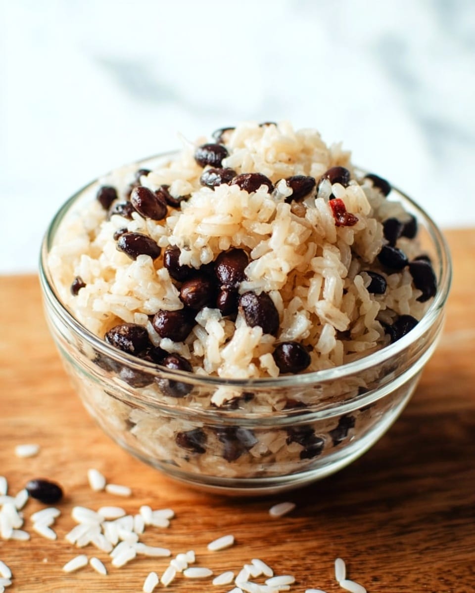 A clear glass bowl sits on a wooden surface filled with two visible layers of cooked rice and black beans mixed together; the rice is light beige with a soft, fluffy texture, and the black beans are dark and glossy. Some grains of white rice are scattered around the bowl on the wooden surface. The background shows a clean, bright white marbled texture. Photo taken with an iphone --ar 4:5 --v 7