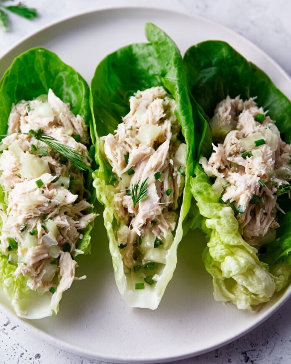 The image shows several small lettuce leaves, each holding a scoop of creamy chicken salad mixed with small bits of herbs, giving a fresh green speckled look on top. The leaves are bright green and crisp, serving as natural cups for the pale beige chicken mixture. They are placed on a white marbled surface with a white wooden cutting board in the background holding more lettuce cups. The front lettuce cup is in clear focus, showing the texture of shredded chicken and fresh herbs, while the others fade softly into the background. photo taken with an iphone --ar 4:5 --v 7