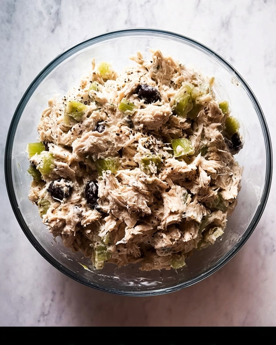 A clear glass bowl filled with a chunky mixture of light beige shredded chicken, mixed with small pieces of green celery and some darker bits that could be raisins or olives, all sprinkled generously with coarse black pepper. The texture looks moist and soft, with visible chunks of both meat and vegetables, sitting on a white marbled surface. The overall look is a hearty, fresh chicken salad with contrasting colors and a rustic, home-made feel. photo taken with an iphone --ar 4:5 --v 7