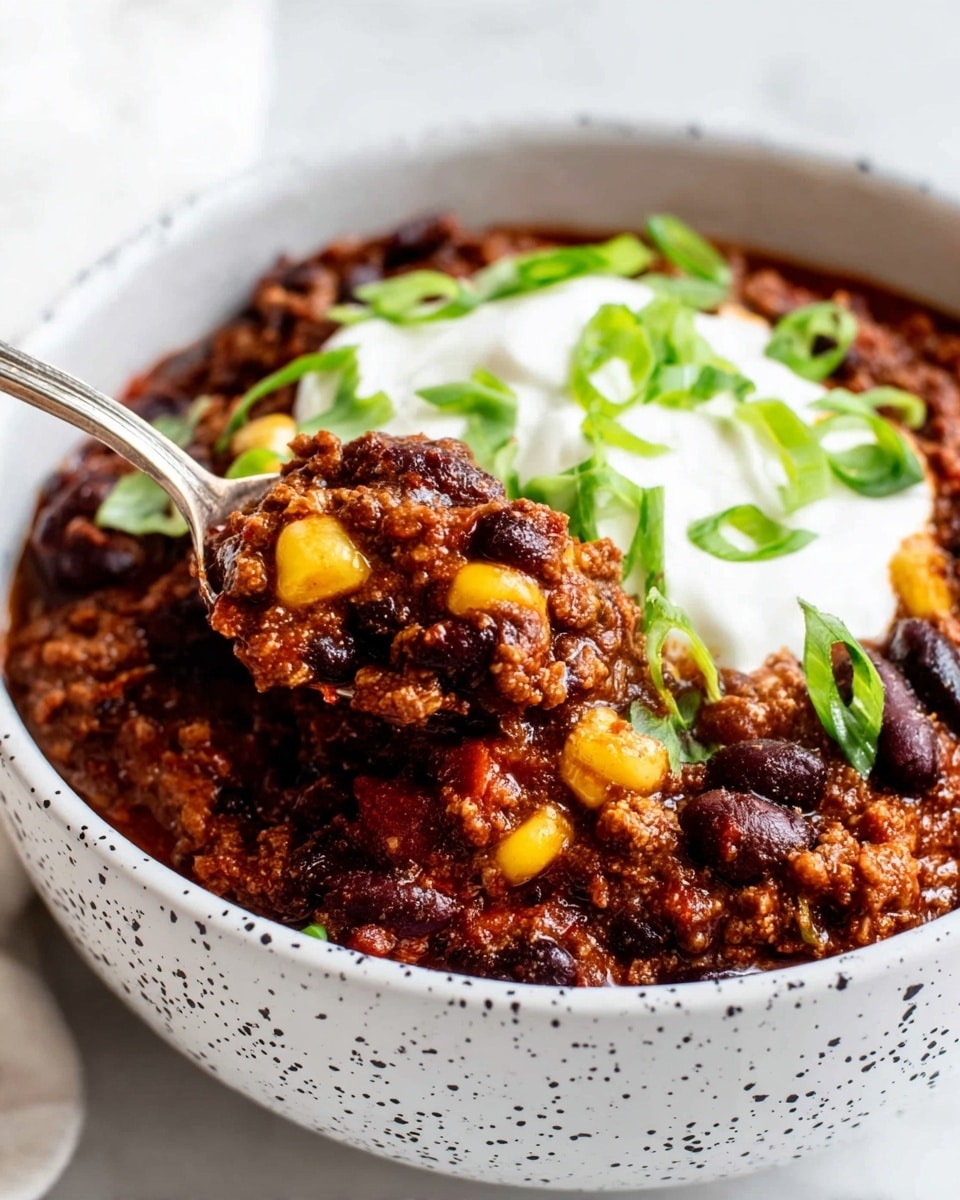 The image shows a white speckled bowl filled with a layered chili dish. The bottom layer consists of dark red kidney beans and black beans mixed with tender ground beef and bright yellow corn kernels, all coated in a rich, deep red sauce. On top, there is a melted layer of golden cheddar cheese partially hidden under a dollop of fluffy white sour cream. The sour cream is garnished with thin slices of fresh green scallions scattered around. The bowl sits on a white marbled surface with a white cloth napkin showing a black grid pattern on the side. photo taken with an iphone --ar 4:5 --v 7