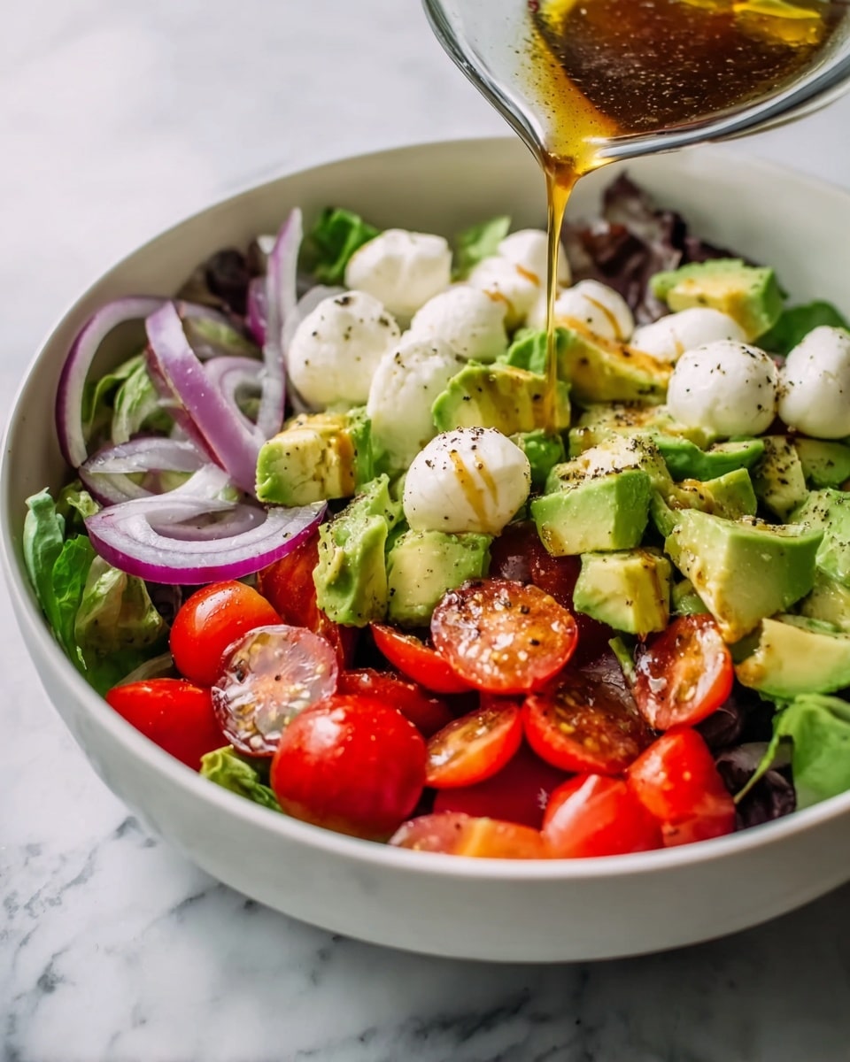 The image shows a fresh salad in a white bowl placed on a white marbled surface. The salad has three main layers: a base of dark green leafy lettuce, a middle layer of chunky green avocado pieces and rings of purple onions, and a top layer scattered with bright red halved cherry tomatoes and small white mozzarella balls. A golden brown dressing is being poured over the salad from a clear glass container, adding a shiny texture. The mix of colors and textures is vibrant and fresh, with visible small black pepper specks on top. photo taken with an iphone --ar 4:5 --v 7