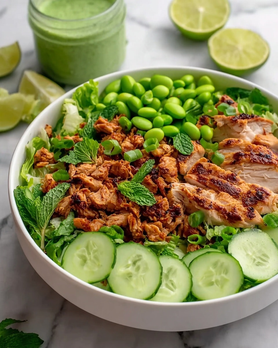 A white bowl filled with a layered salad starts with a base of green leafy lettuce. On top of this are round, light green cucumber slices arranged mostly around the sides. Scattered across the salad are small pieces of browned grilled chicken with a slightly charred texture. There are also bright green edamame beans and chopped green onions sprinkled on top. The dish is garnished with some fresh green mint leaves. In the background, there are slices of lime and a glass jar with a green sauce, all set on a white marbled surface. photo taken with an iphone --ar 4:5 --v 7
