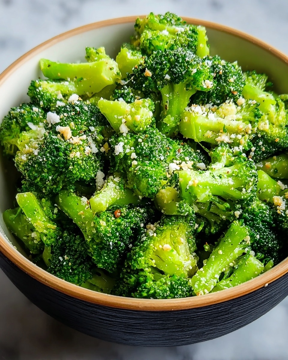 A close-up view of a bowl filled with bright green broccoli florets that are cooked and lightly seasoned, topped with small white and light beige grated cheese or crumb bits scattered evenly over the broccoli. The broccoli is fresh and moist, showing clear details of the tiny buds and stems. The bowl is white on the inside with a dark outer rim, placed on a surface with a white marbled texture. photo taken with an iphone --ar 4:5 --v 7