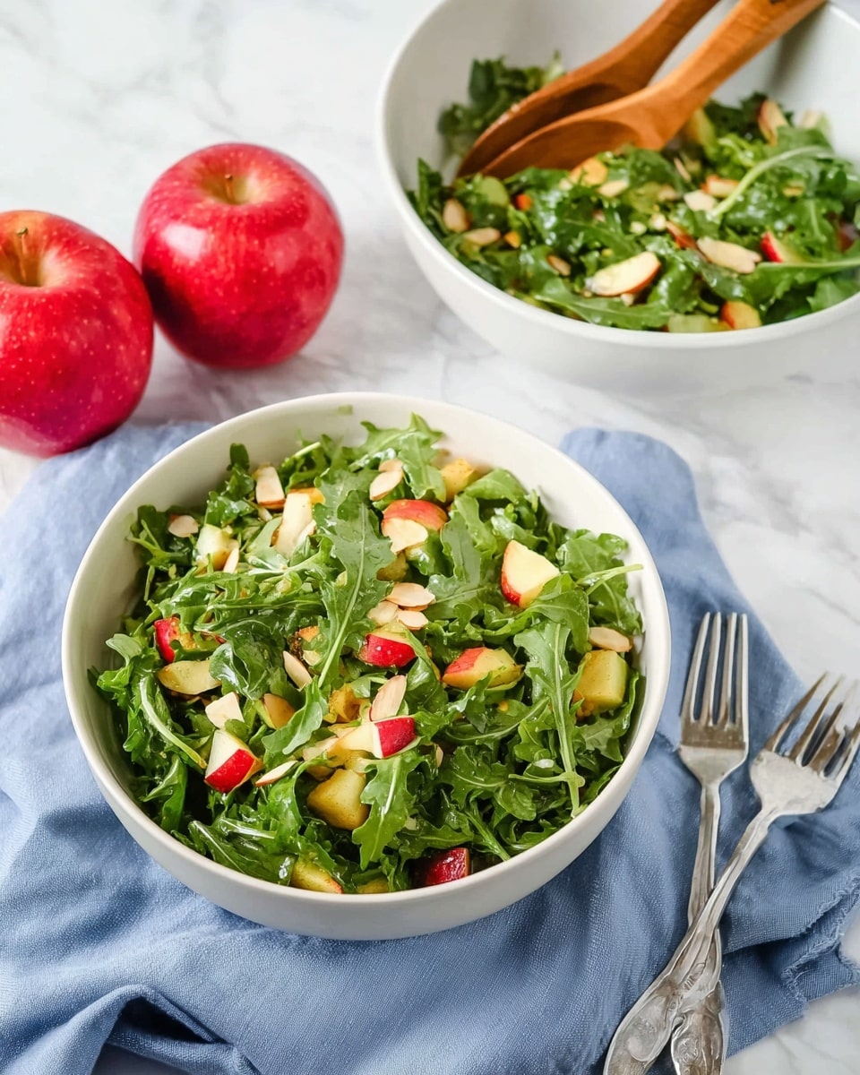 A fresh salad is shown in a white bowl filled with deep green arugula leaves mixed with small, irregular chunks of red and yellow apple pieces scattered throughout. Thin, light brown almond slices are sprinkled on top, adding a crunchy texture contrast. Another white bowl containing the same salad mixture with wooden salad tongs rests in the background on a white marbled surface. Two shiny red apples sit beside the bowls, and a light blue cloth napkin with a silver fork lies next to the salad bowl in the foreground. Photo taken with an iphone --ar 4:5 --v 7
