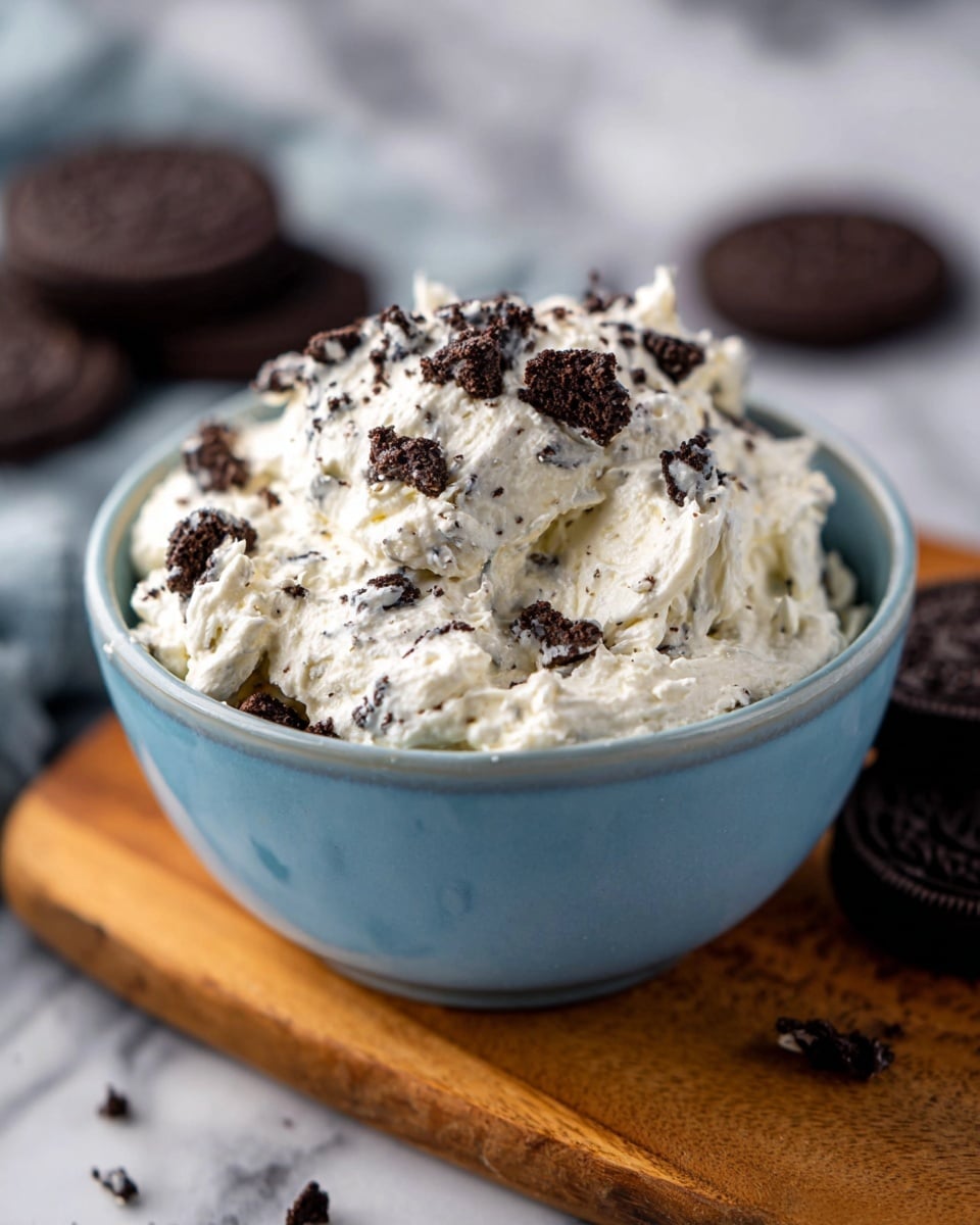 A close-up view of a light blue bowl filled with a creamy white mixture that has a fluffy texture, topped with scattered small chunks of dark chocolate cookies. The bowl sits on a wooden board, with two whole chocolate cookies blurred in the background. The scene is set on a white marbled surface, highlighting the contrast between the creamy mixture and the dark cookie pieces. Photo taken with an iphone --ar 4:5 --v 7