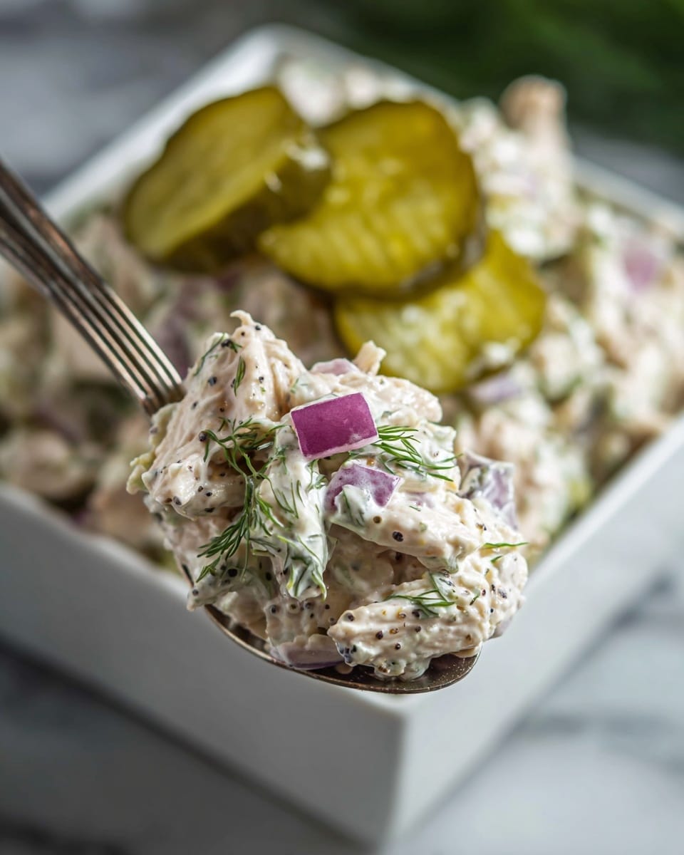A close-up view of a spoonful of creamy chicken salad with visible layers including tender white shredded chicken mixed evenly with white mayonnaise dressing, speckled with black pepper, small pieces of purple-red onion, and green dill sprinkled on top. The background shows a white square bowl filled with more of the same salad, garnished with several sliced pickles that are light green with a slightly translucent texture. The setting rests on a white marbled surface. photo taken with an iphone --ar 4:5 --v 7