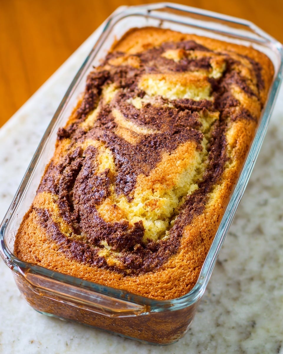 A loaf of marble cake with two layers baked in a clear glass loaf pan, resting on a white marbled texture. The cake has a golden brown base with swirls of dark brown chocolate mixed across the top, creating a marbled pattern. The surface texture is slightly cracked and crispy, showing a mix of smooth and crumbly areas with visible color contrast between the light yellowish cake and the dark chocolate swirls. Photo taken with an iphone --ar 4:5 --v 7