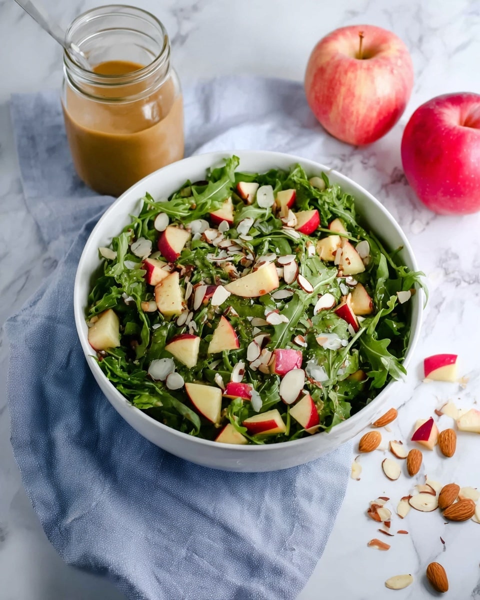A white bowl filled with a fresh salad showing three clear layers: a base layer of dark green arugula leaves with a rough texture, scattered with light beige almond slices as the middle layer, and topped with small chunks of red and cream-colored apple pieces adding bright color contrast. The bowl sits on a light blue cloth, placed on a white marbled surface. Two whole apples, one red and one pinkish-red, are on the right side near the bowl, along with some scattered almond slices. A glass jar containing light brown dressing is positioned above the bowl. Photo taken with an iphone --ar 4:5 --v 7