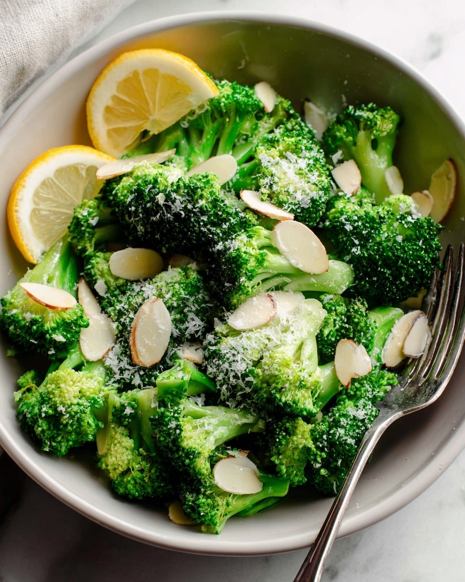 The dish shows several bright green broccoli florets arranged in a white bowl. On top of the broccoli, there are thin slices of pale almond pieces scattered evenly. Light sprinkles of grated white cheese or powdery topping cover some broccoli parts. Two lemon wedges with pale yellow and white flesh rest on the broccoli at the back left of the bowl. A metal fork is placed inside the bowl on the right side, partly visible. The bowl is set on a white marbled surface. photo taken with an iphone --ar 4:5 --v 7