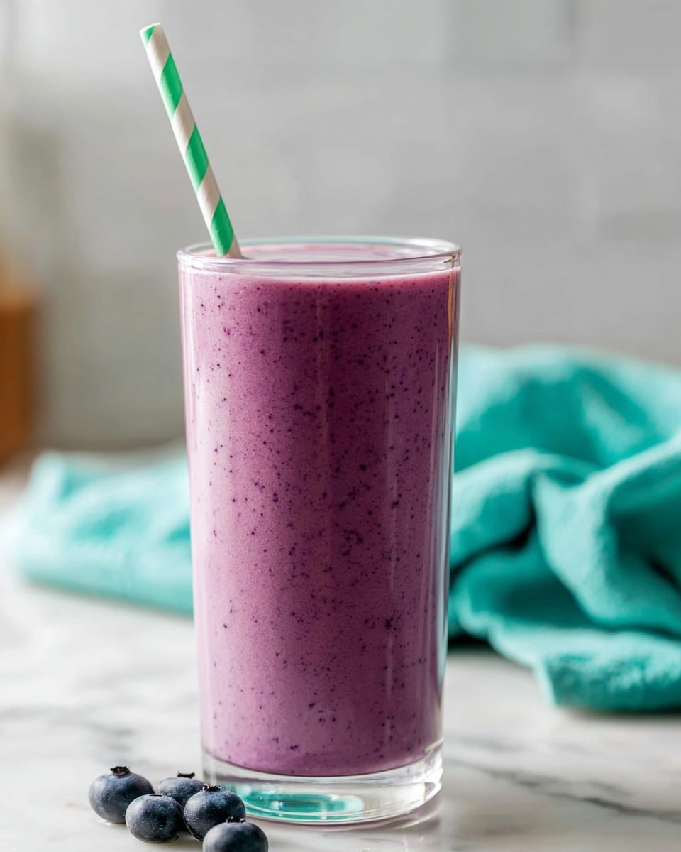 A tall clear glass filled with a thick, creamy purple smoothie that has tiny dark specks throughout, giving it a rich texture; a green and white striped paper straw is inserted at the top, poking out slightly at an angle. The glass is set on a white marbled surface, with part of a turquoise cloth blurred in the background and a few fresh blueberries on the surface near the glass. The photo has soft, natural lighting and a shallow depth of field that keeps the smoothie sharp and the background smooth. photo taken with an iphone --ar 4:5 --v 7