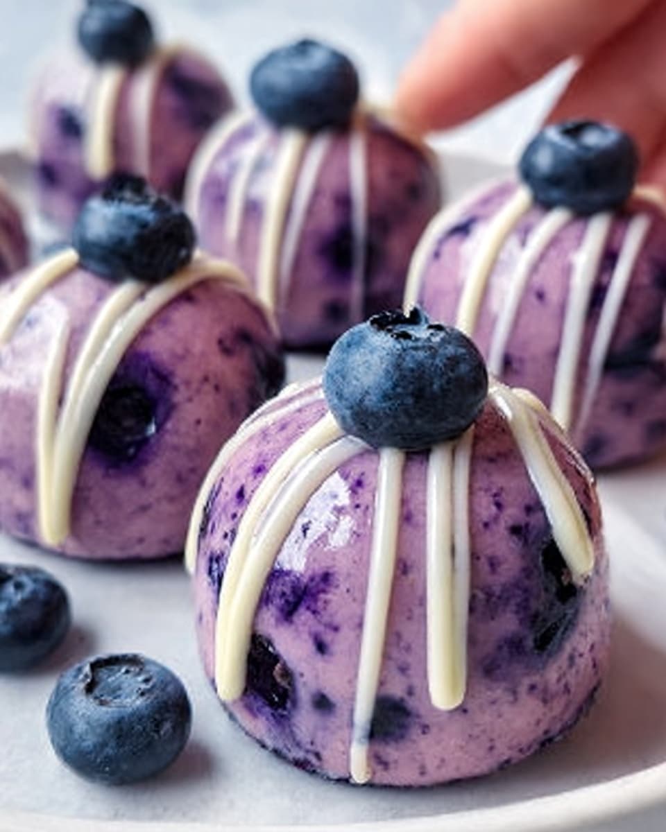 The image shows small round desserts with a smooth, shiny purple outer layer that looks creamy and speckled with darker blueberry bits. Each dessert is topped with two fresh blueberries and decorated with three white icing stripes running from the top to the sides, creating a neat pattern. The desserts are placed closely together on a white plate set on a white marbled surface, and a woman's hand is gently touching one of them. The lighting highlights the glossy texture and vibrant purple color of the treats, making them look fresh and delicious. photo taken with an iphone --ar 4:5 --v 7