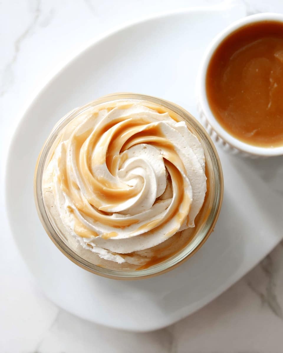 A clear glass jar filled with layers of creamy dessert is shown from above, topped with a swirl of soft whipped cream mixed with caramel-colored sauce, creating a two-tone spiral pattern. The jar sits on a white plate with a smooth, white marbled texture surface beneath. To the top right of the jar, there is a white bowl filled with a smooth caramel sauce. The soft, light textures and warm creamy tones contrast against the bright, clean white background. Photo taken with an iphone --ar 4:5 --v 7