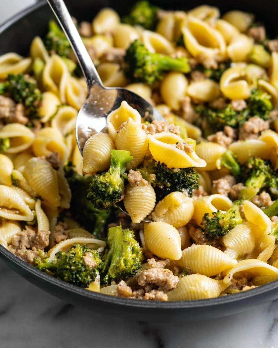 A close-up view of a pasta dish in a black pan on a white marbled surface, showing three main layers mixed together: small pale yellow shell pasta with a soft and smooth texture, pieces of light brown cooked ground meat with a slightly crumbly look, and small green broccoli florets with a fresh and bumpy surface. A shiny silver spoon is scooping the mix from the pan, reflecting the colors and textures of the food. photo taken with an iphone --ar 4:5 --v 7