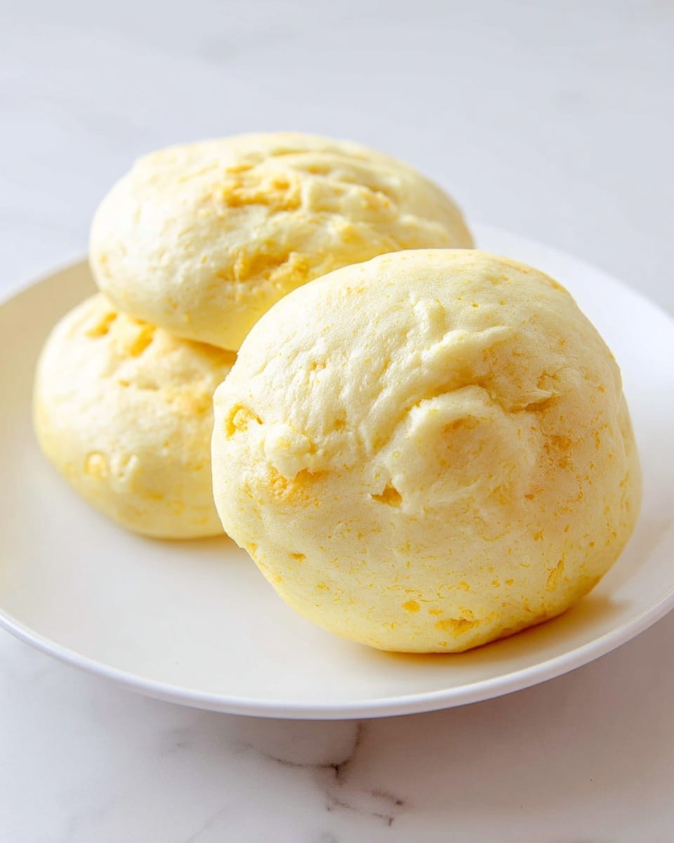 Two yellowish steamed buns with a soft, slightly textured surface sit on a smooth white plate. The buns have a few lighter and darker spots, showing a gentle crumbly texture and small folds on the top. One bun is in the front right side, fully visible, while the other two are stacked slightly behind on the left, giving a sense of depth. The plate rests on a white marbled surface, adding brightness to the image. photo taken with an iphone --ar 4:5 --v 7