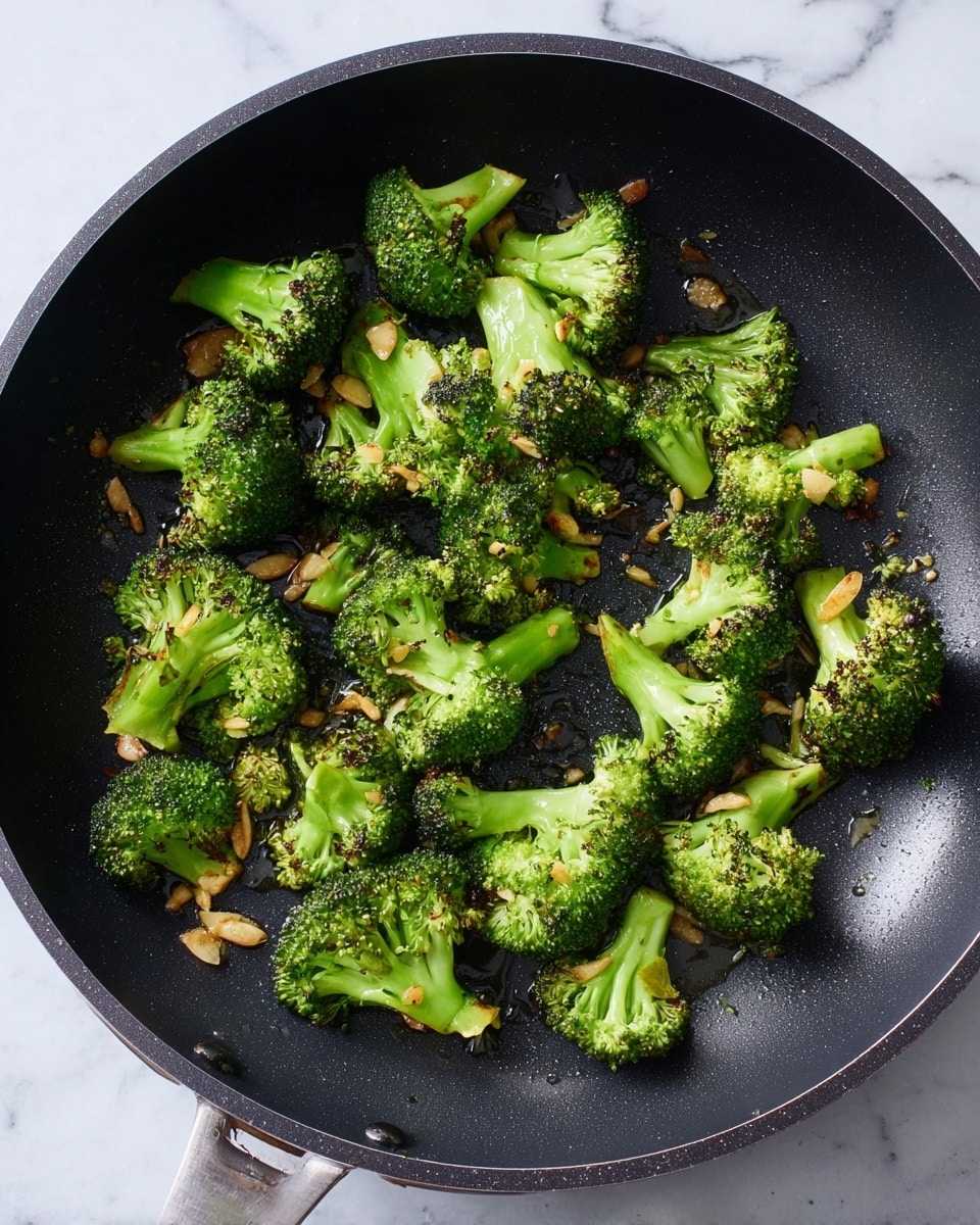 A black cooking pan holds several pieces of cooked broccoli florets scattered evenly across its surface; the broccoli is bright green with some darker charred spots showing where it was grilled or sauteed. Thin, golden-brown slices of garlic are mixed in among the broccoli, adding texture and color contrast. Tiny droplets of oil glisten on the broccoli and pan, giving a fresh and oily look. The pan sits on a surface with a white marbled texture. photo taken with an iphone --ar 4:5 --v 7