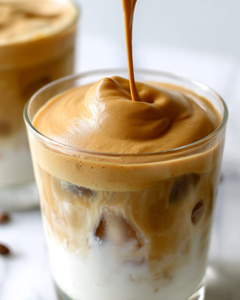 A clear glass cup filled with a three-layered iced drink sits on a white marbled surface. The bottom layer is white milk with visible ice cubes, the middle layer is a slightly light brown coffee mix blending softly into the milk, and the top layer is a thick, smooth, and creamy caramel brown frothy foam being poured into the cup, creating a soft peak. A second similar drink blurred in the background adds depth to the scene. Photo taken with an iphone --ar 4:5 --v 7