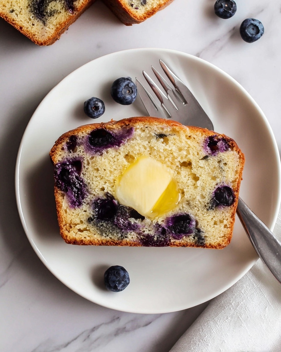 A white plate holds a single slice of blueberry bread with a golden-brown crust and a light, spongy interior filled with whole blueberries that give purple stains inside the bread. On top of the bread slice, there is a melting pat of yellow butter in the center. To the right side of the plate rests a silver fork. Around the plate on a white marbled surface, there are a few loose blueberries and two other slices of the same bread partially visible in the upper left corner. photo taken with an iphone --ar 4:5 --v 7