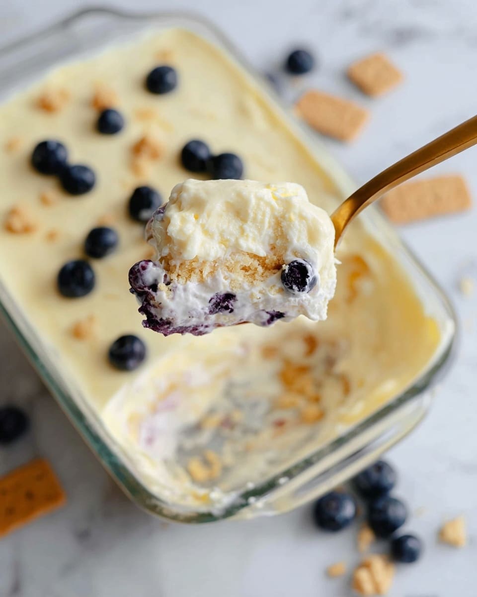 A close-up view of a creamy dessert served in a clear glass rectangular dish placed on a white marbled surface. The dessert has a soft, pale yellow top layer with a smooth texture, dotted with whole dark blueberries. A golden spoon is shown holding a scoop of the dessert above the dish, revealing two layers: the top fluffy cream layer with a light, smooth texture and embedded blueberries, and a bottom layer with a crumbly, light brown texture, likely crushed cookies or crust. Scattered blueberries and bits of broken cookies are blurred in the background. Photo taken with an iphone --ar 4:5 --v 7