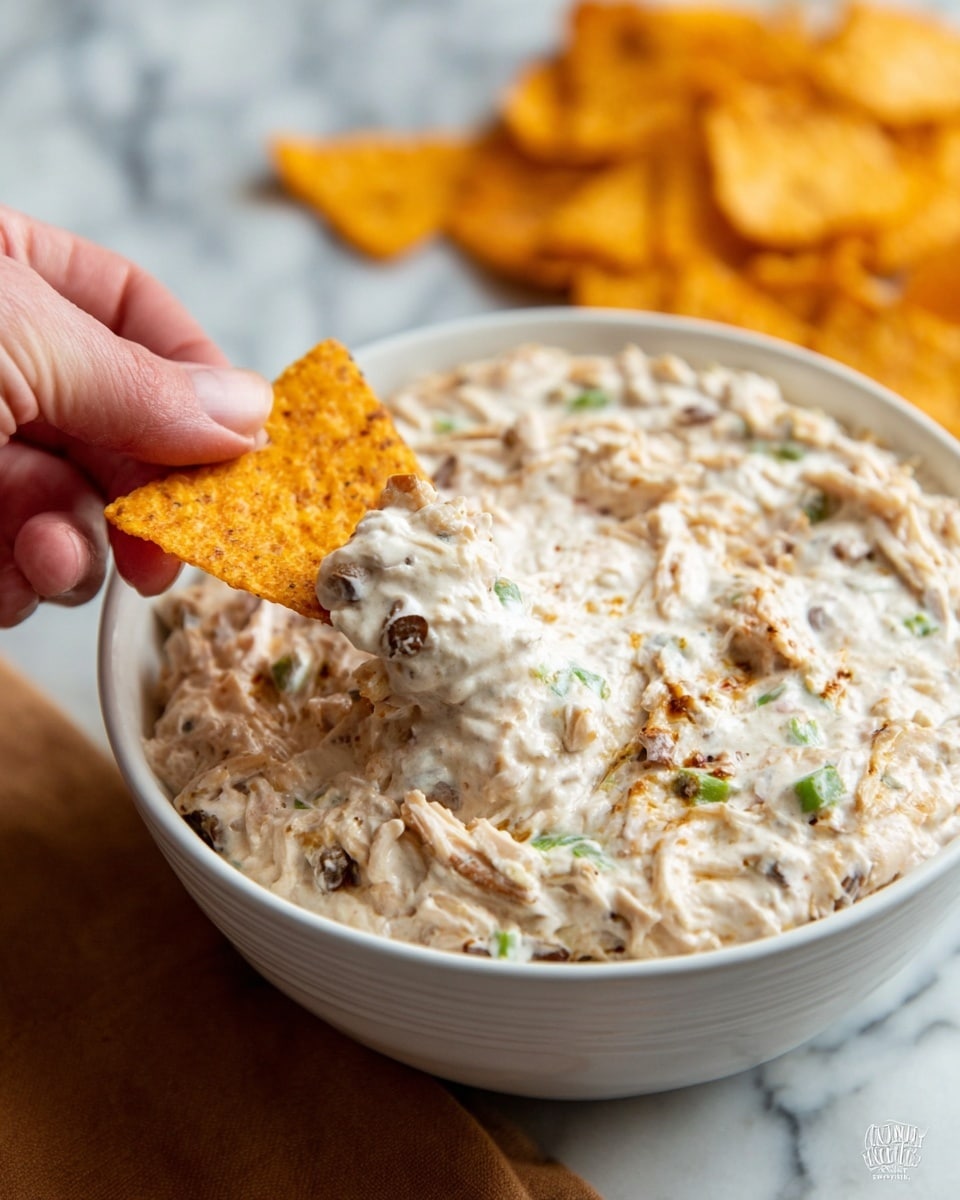 A close-up view of a white bowl filled with creamy chicken dip that has visible small green and brown bits mixed in. The dip looks thick and smooth with shredded chicken pieces spread all through it. A crispy orange-brown chip, held by a woman's hand on the left, is dipped halfway into the dip, showing a thick layer of the creamy mix covering its tip. The bowl sits on a white marbled surface, and extra chips are scattered in soft focus behind the bowl. Photo taken with an iphone --ar 4:5 --v 7