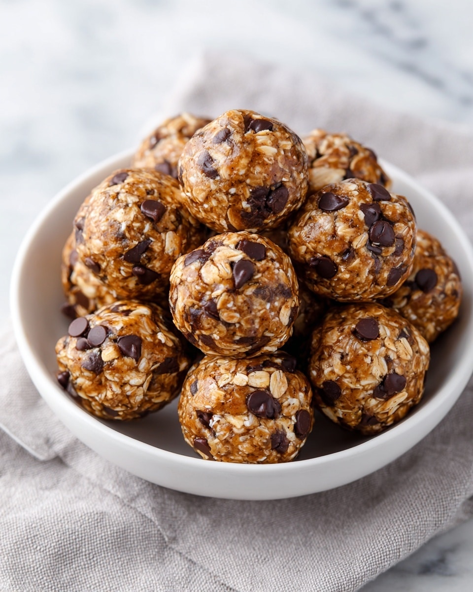 A pile of round energy balls in a shallow white plate, each ball showing visible oats mixed with dark brown chocolate and studded with dark chocolate chips, creating a textured look. The balls are dense and packed together, with the oats adding a light beige contrast to the dark chocolate base. The plate sits on a soft grey cloth over a white marbled surface, and warm natural light highlights the shiny and rich texture of the chocolate balls. photo taken with an iphone --ar 4:5 --v 7