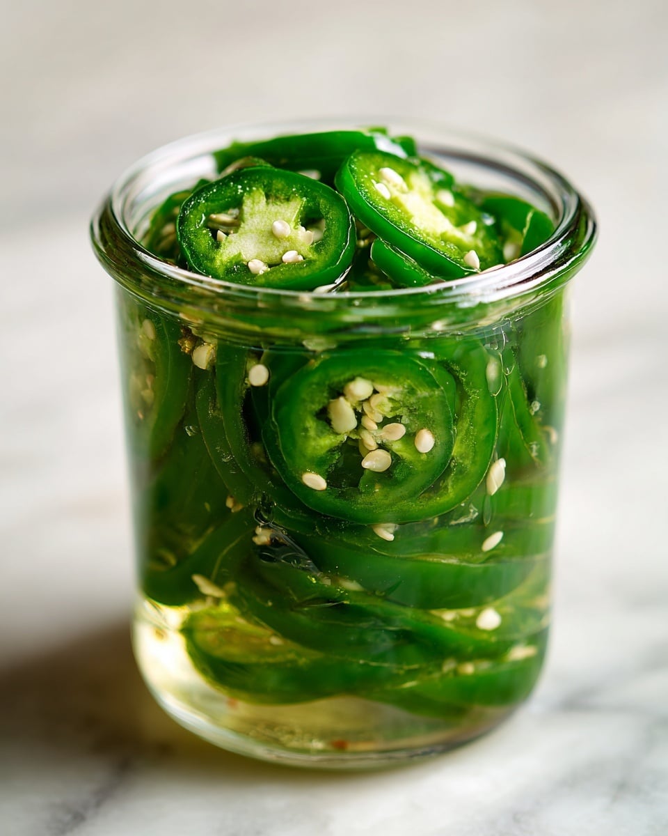 A clear glass jar filled with several layers of green jalapeño slices, each slice showing white seeds and a shiny, wet texture indicating they are pickled. The jalapeño slices are tightly packed inside the jar, with some seeds floating in the clear brine liquid that fills the spaces between the slices. The jar is placed on a white marbled surface, and the background is softly blurred to focus on the vibrant green peppers inside the jar. photo taken with an iphone --ar 4:5 --v 7