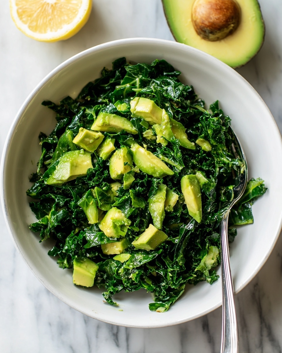 A white bowl filled with a fresh kale salad mixed with chunks of light green avocado pieces scattered evenly on top and throughout the dark green kale leaves, sprinkled with black pepper and small bits of lemon zest, with a silver spoon on the right side inside the bowl. Next to the bowl on the white marbled surface are a half avocado with its seed removed and a small lemon wedge. Photo taken with an iphone --ar 4:5 --v 7