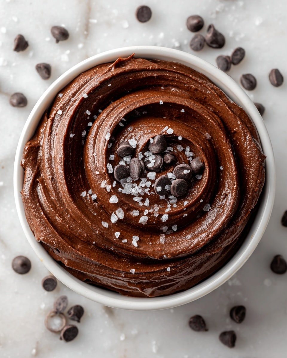 A bowl filled with a smooth, dark brown chocolate mousse swirled in a spiral pattern, topped with a small pile of dark chocolate chips in the center and scattered coarse sea salt flakes across the surface. The bowl is white, resting on a white marbled texture with a grey cloth nearby. In the background, there are small bowls with more coarse salt and sliced red strawberries slightly blurred. Photo taken with an iphone --ar 4:5 --v 7