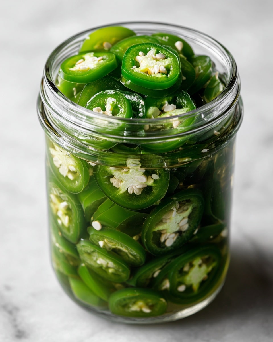 A clear glass jar filled with multiple layers of green jalapeño slices, each slice showing a glossy texture with visible seeds and pale green inner flesh, all soaked in a translucent brine that fills the jar almost to the top. The jar sits on a white marbled surface, and the light reflects slightly on the glass, highlighting the shiny, wet jalapeños inside. photo taken with an iphone --ar 4:5 --v 7