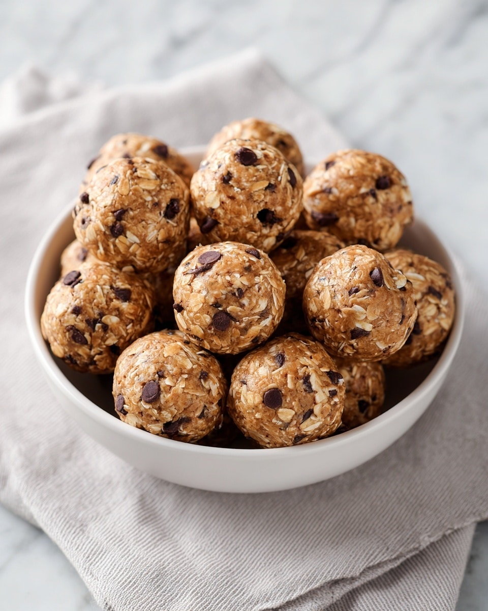 A white bowl holds a pile of round snack balls, each made from oats, chocolate chips, and a sticky brown mixture that binds them together. The balls are tightly packed, with the oats showing light beige flecks and the chocolate chips as dark brown spots scattered on the surface. The texture looks chewy with bits of oats visible and shiny chocolate spots. The bowl sits on a light gray cloth, and the background is a white marbled texture. photo taken with an iphone --ar 4:5 --v 7