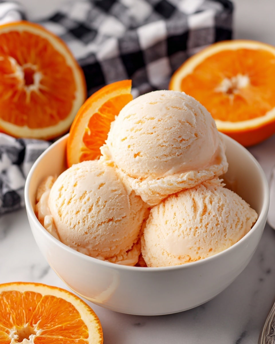 A white bowl filled with three scoops of pale orange ice cream with a soft, creamy texture, topped with a small slice of bright orange with visible pulp placed on the rim of the bowl; in the background, two orange halves show their juicy interior, all set on a white marbled surface with a blurred black and white checkered cloth behind. photo taken with an iphone --ar 4:5 --v 7