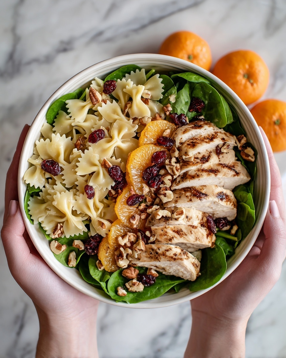 A white bowl filled with a layered salad starting with a base of fresh green spinach leaves, topped with a layer of cooked bow-tie pasta that is pale yellow. Scattered on top are pieces of grilled chicken with char marks, along with bright orange segments and dark red dried cranberries. Small round nuts are sprinkled throughout, adding texture and light brown color to the dish. A woman's hands hold the bowl on each side, and the background is a white marbled surface with some green leaves and orange fruits nearby. photo taken with an iphone --ar 4:5 --v 7