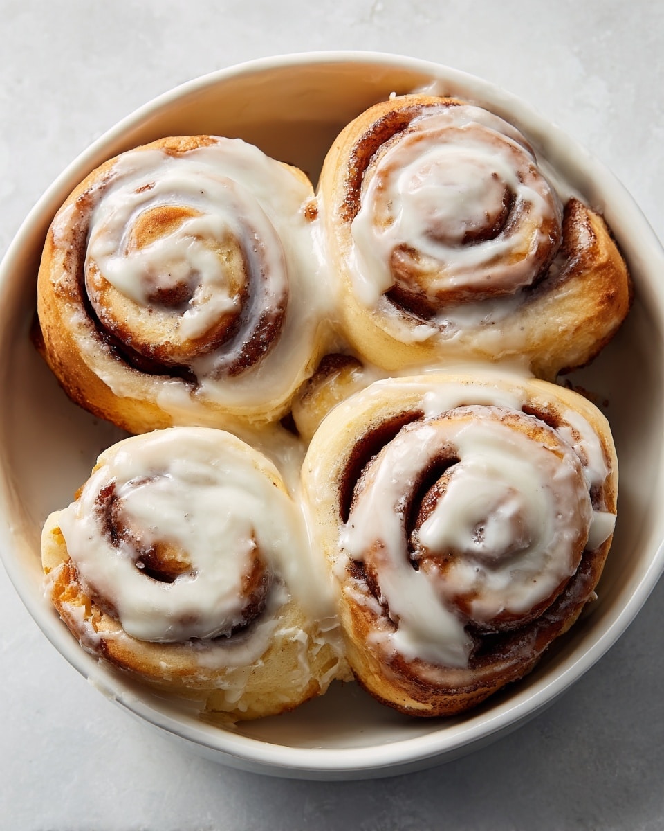 A white bowl filled with five cinnamon rolls, each with a slightly golden-brown outer layer and swirls of dark cinnamon inside, topped with smooth white icing that is spread more thickly on some rolls and lightly on others, showing shiny and soft textures. The rolls are arranged closely together, slightly touching, and the white marbled surface background adds a clean and bright look to the image. Photo taken with an iphone --ar 4:5 --v 7