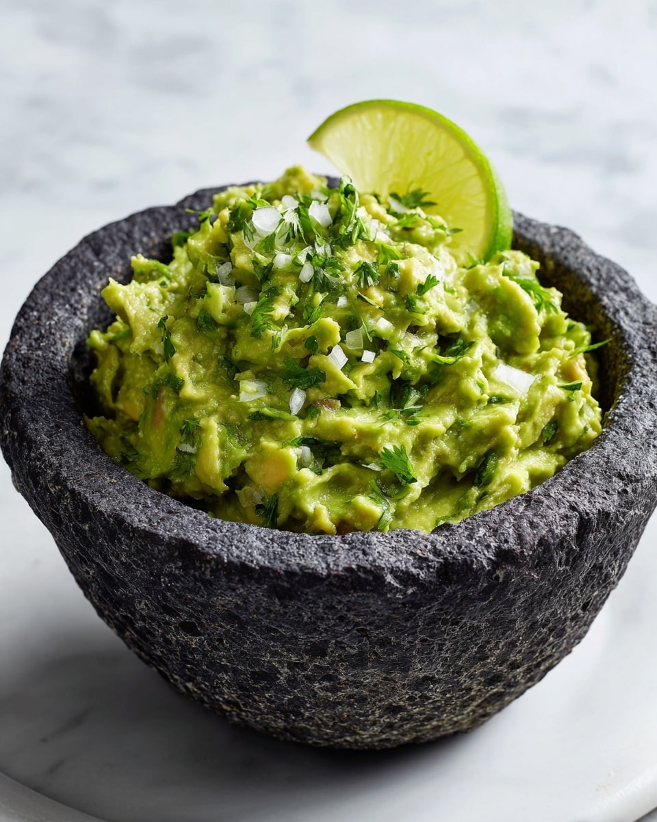 A close-up view of guacamole in a dark gray, textured stone bowl, showing a thick mix of mashed and chunky avocado pieces in bright green with bits of white onion and fresh chopped cilantro sprinkled throughout; a wedge of lime with a vibrant yellow-green rind rests on the right side inside the bowl. The bowl is placed on a surface with a white marbled texture, with a part of a white plate visible in the top left corner and a small visible edge of a bowl with crispy chips at the bottom left. Photo taken with an iphone --ar 4:5 --v 7