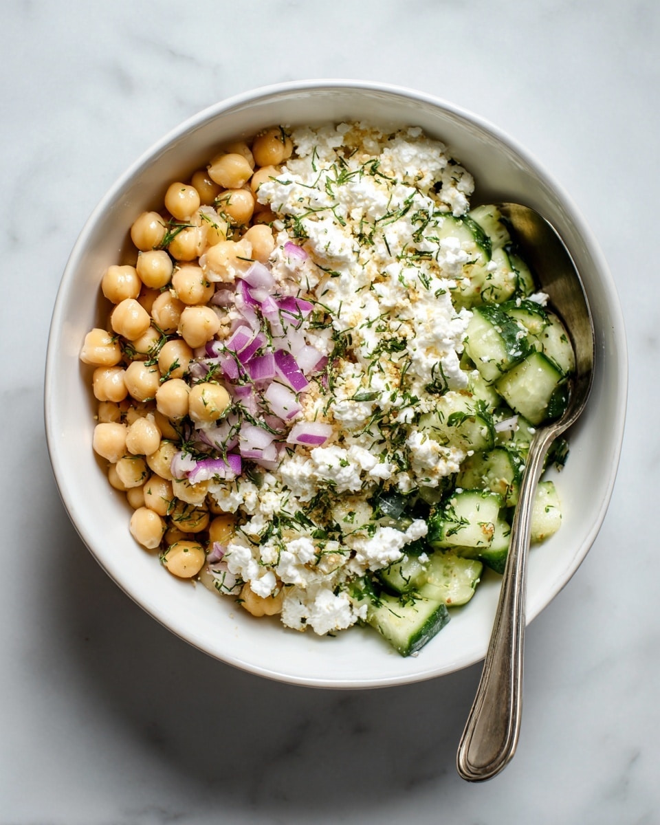 A white bowl filled with a chickpea salad made of three main layers: a base layer of small white cottage cheese curds mixed with chickpeas, a middle layer of light green cucumber pieces, and scattered tiny bits of purple onion and chopped green herbs evenly spread throughout. The salad is mixed well, giving a creamy and chunky texture. A silver spoon is placed inside the bowl on the right side. The bowl sits on a white marbled surface. photo taken with an iphone --ar 4:5 --v 7