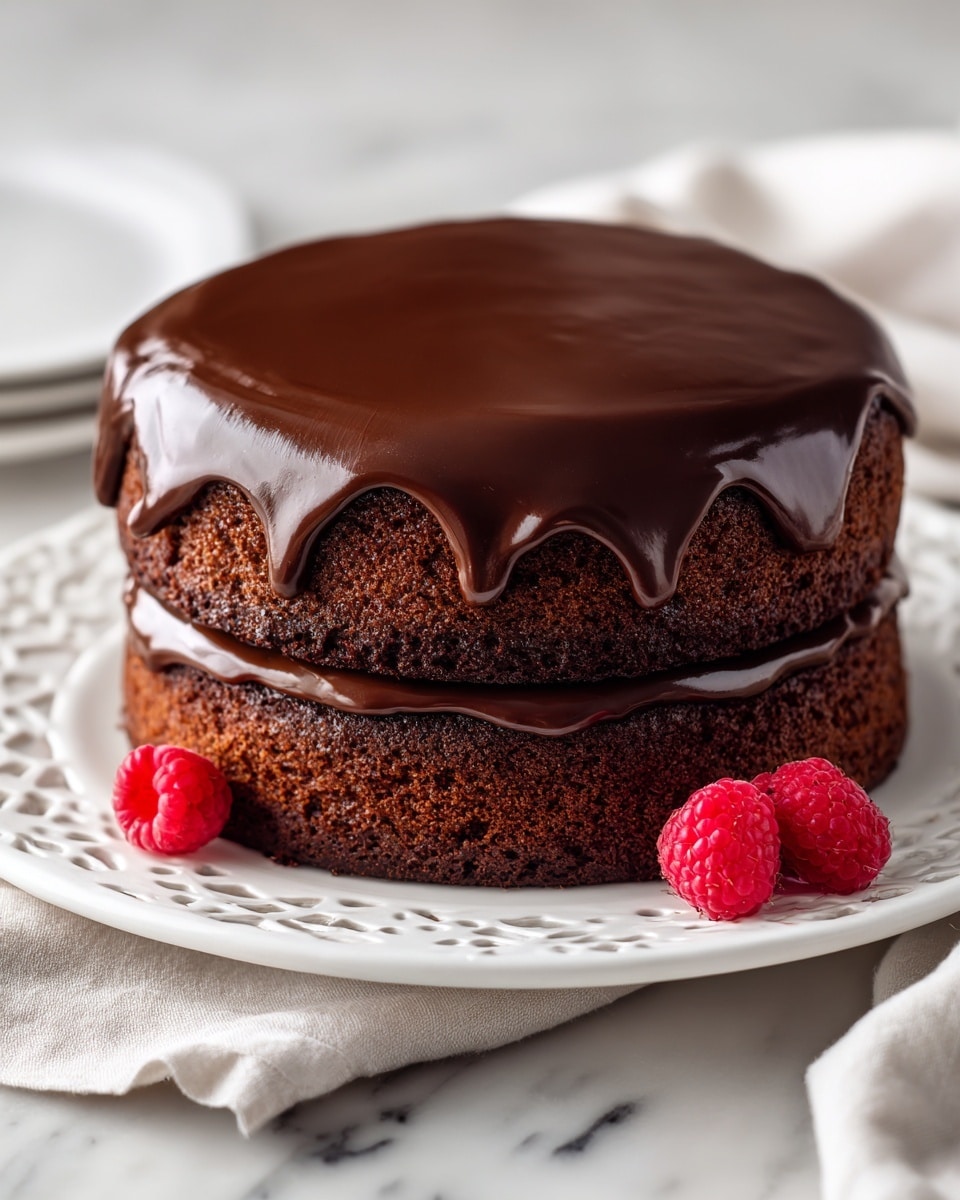 A two-layer chocolate cake sits on a white plate with delicate cut-out patterns. The bottom layer is thick, dark brown, and has a moist texture. Between the layers is a smooth, shiny chocolate frosting that spreads evenly. On top, a thick layer of glossy chocolate ganache drips slightly over the sides, adding richness. Two fresh red raspberries sit next to the cake on the plate, adding a pop of color. The setting is on a white marbled surface with a soft cloth underneath the plate. Photo taken with an iphone --ar 4:5 --v 7
