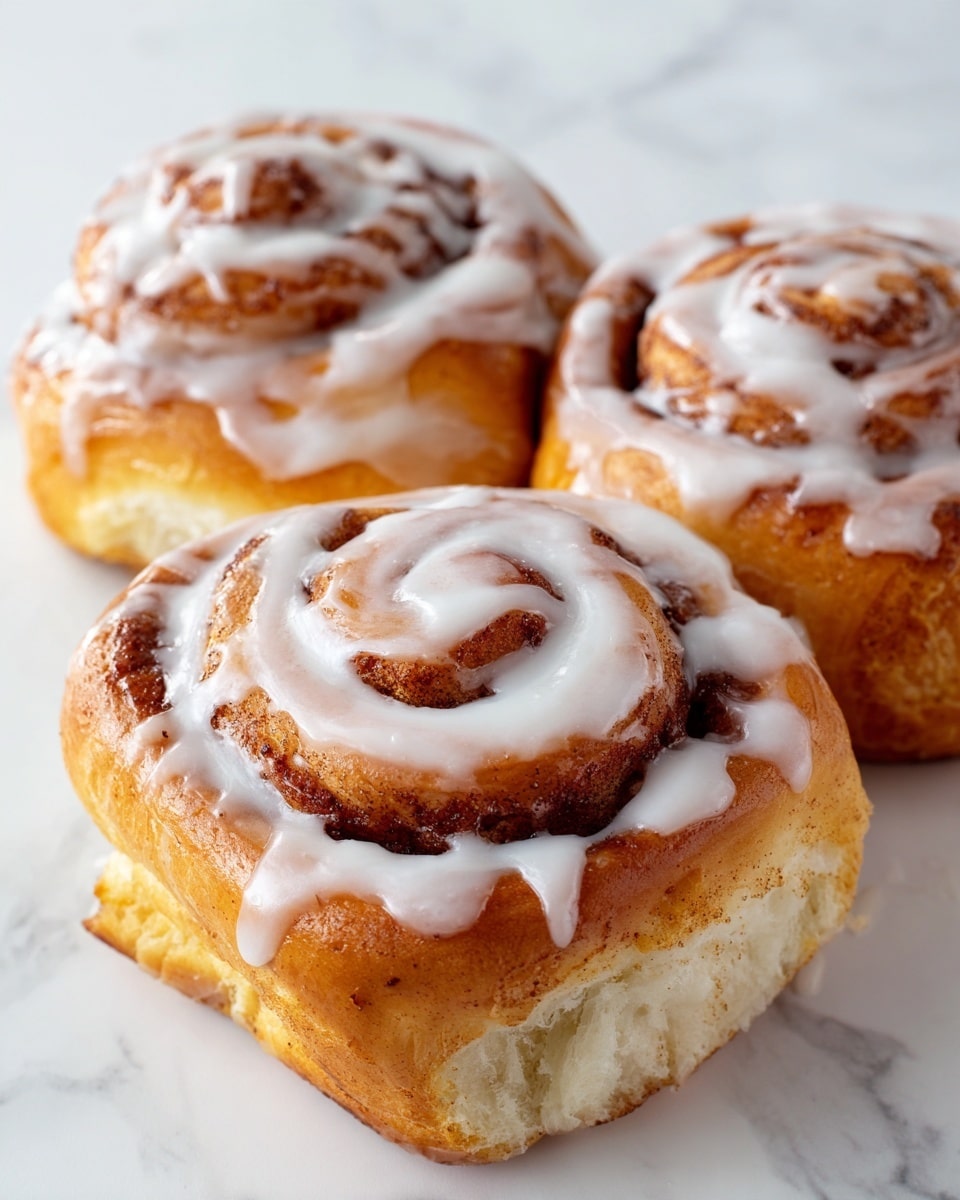 The image shows three cinnamon rolls on a white plate placed on a white marbled surface. Each cinnamon roll has a golden-brown outer crust that looks soft and slightly shiny. The top of each roll is covered with a smooth, glossy layer of white icing that melts down the sides, with a small swirl of extra icing in the center. The rolls have visible spiral layers inside, revealing a cinnamon-sugar mixture with a light brown color. In the background, there is a blurred yellow lemon adding a soft touch of color. photo taken with an iphone --ar 4:5 --v 7