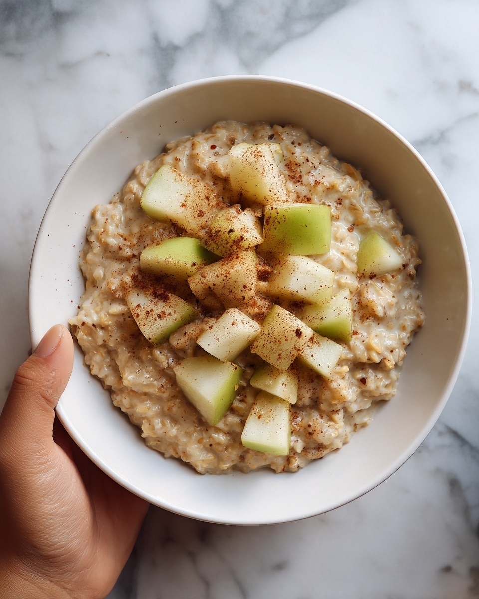 The image shows a white bowl filled with creamy oatmeal mixed with small chunks of soft, light brown spiced pears. The oatmeal texture looks thick and smooth with visible bits of pear scattered throughout, adding a juicy, slightly glossy contrast. A woman's hand is holding the bowl from the side, gently lifting it. The background and surface are white marbled texture, giving a clean and bright look. photo taken with an iphone --ar 4:5 --v 7