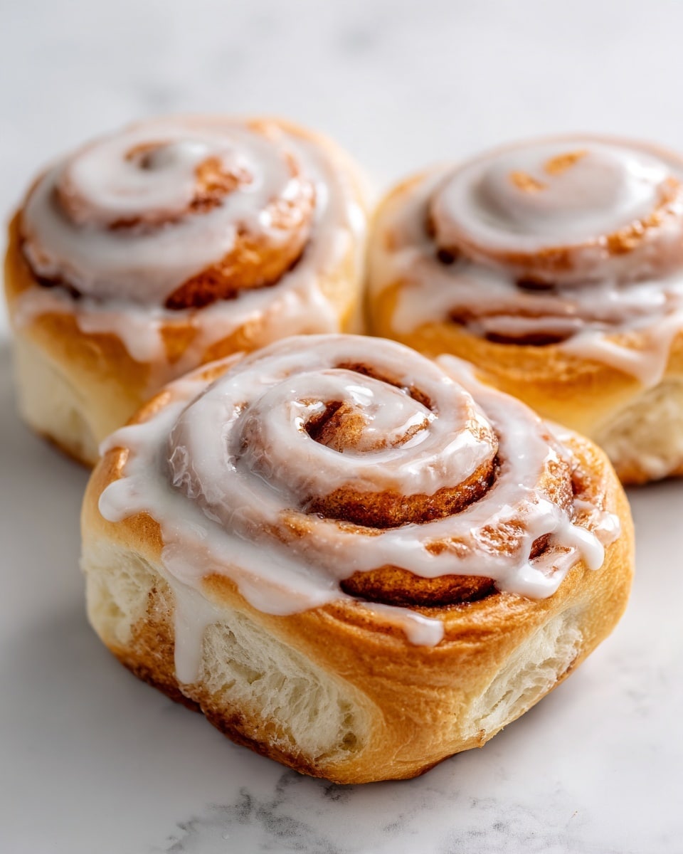 The image shows three warm cinnamon rolls with thick white icing drizzled over the top, creating a glossy texture. Each roll has visible spiral layers of soft brown dough with a slightly golden crust, and the icing pools slightly in the center and along the ridges of the swirls. The cinnamon inside gives the rolls a speckled look within the layers. They are placed closely together on a white surface with a white marbled texture in the background, which adds a clean and bright feel to the image. Photo taken with an iphone --ar 4:5 --v 7